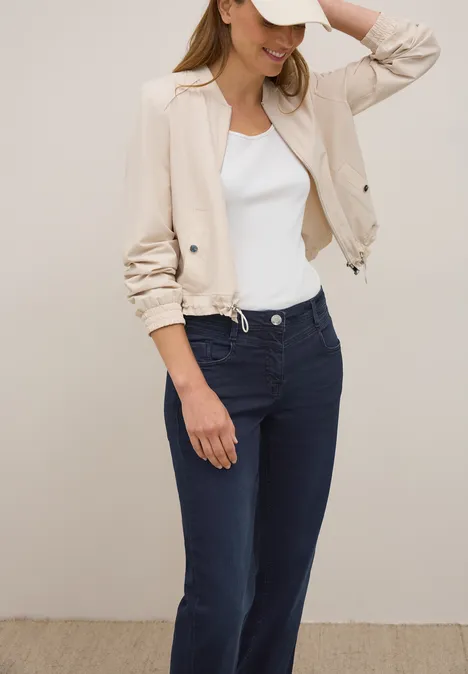Frau mit beiger Jacke, weißem Top, blauer Hose und Basecap, lächelt.